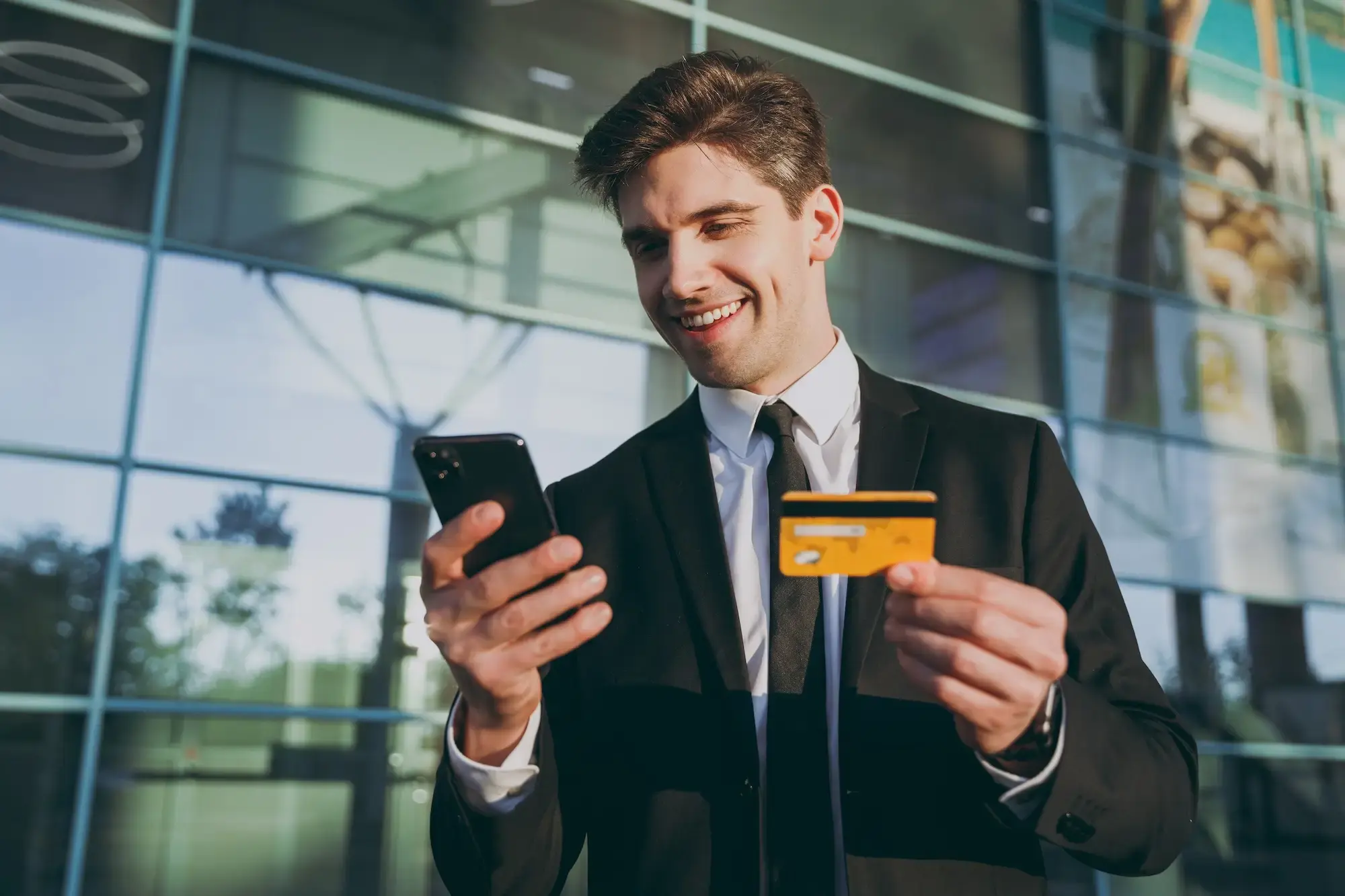 A man in a black suit using a mobile phone and holding a banking card.